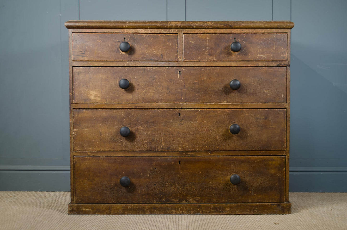 A vintage wooden five-drawer chest with dark round knobs, set against a dark blue paneled wall and a light beige floor.