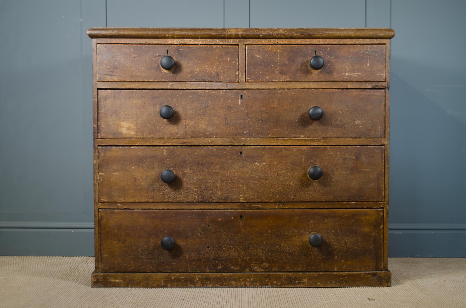 
A vintage wooden five-drawer chest with dark round knobs, set against a dark blue paneled wall and a light beige floor.