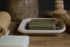 Green bar of soap on a white soap dish with a brush and loofah in the background.