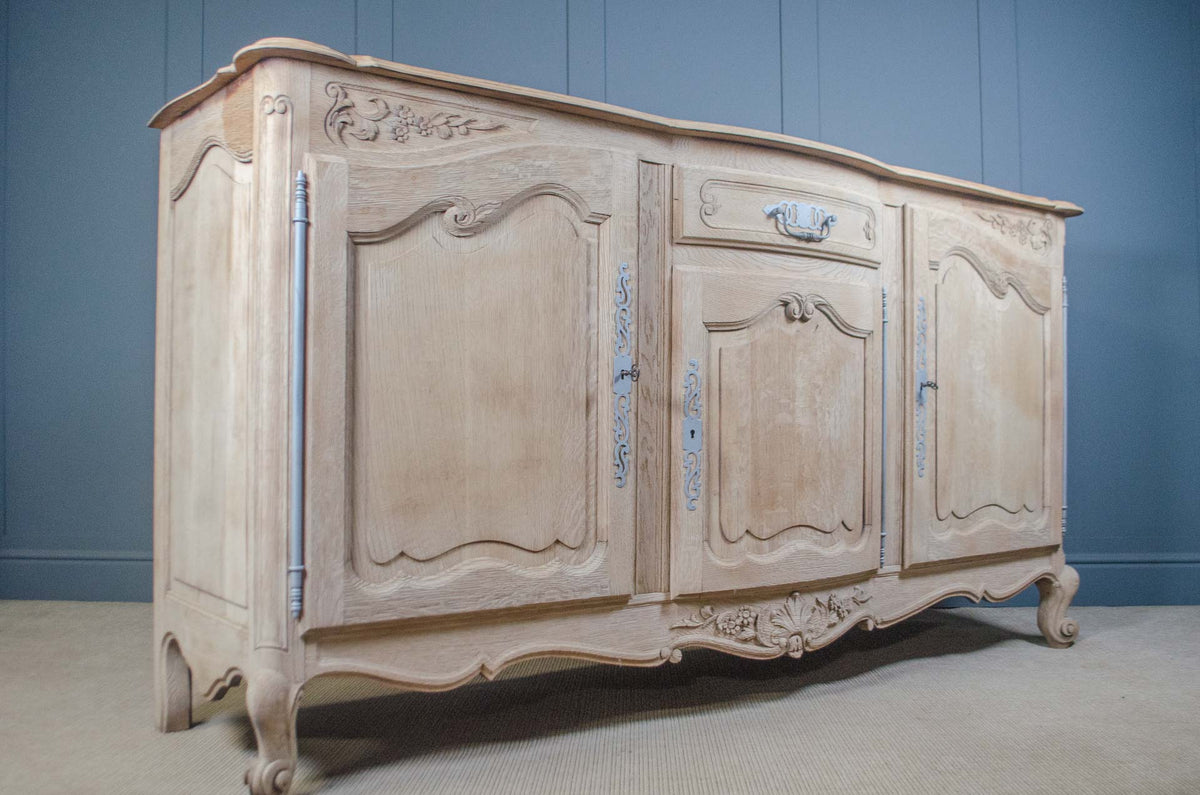 
A distressed French-style sideboard cabinet with carved details, featuring a light beige/cream finish with subtle blue painted accents on the drawer pulls and trim, set against a blue paneled wall.