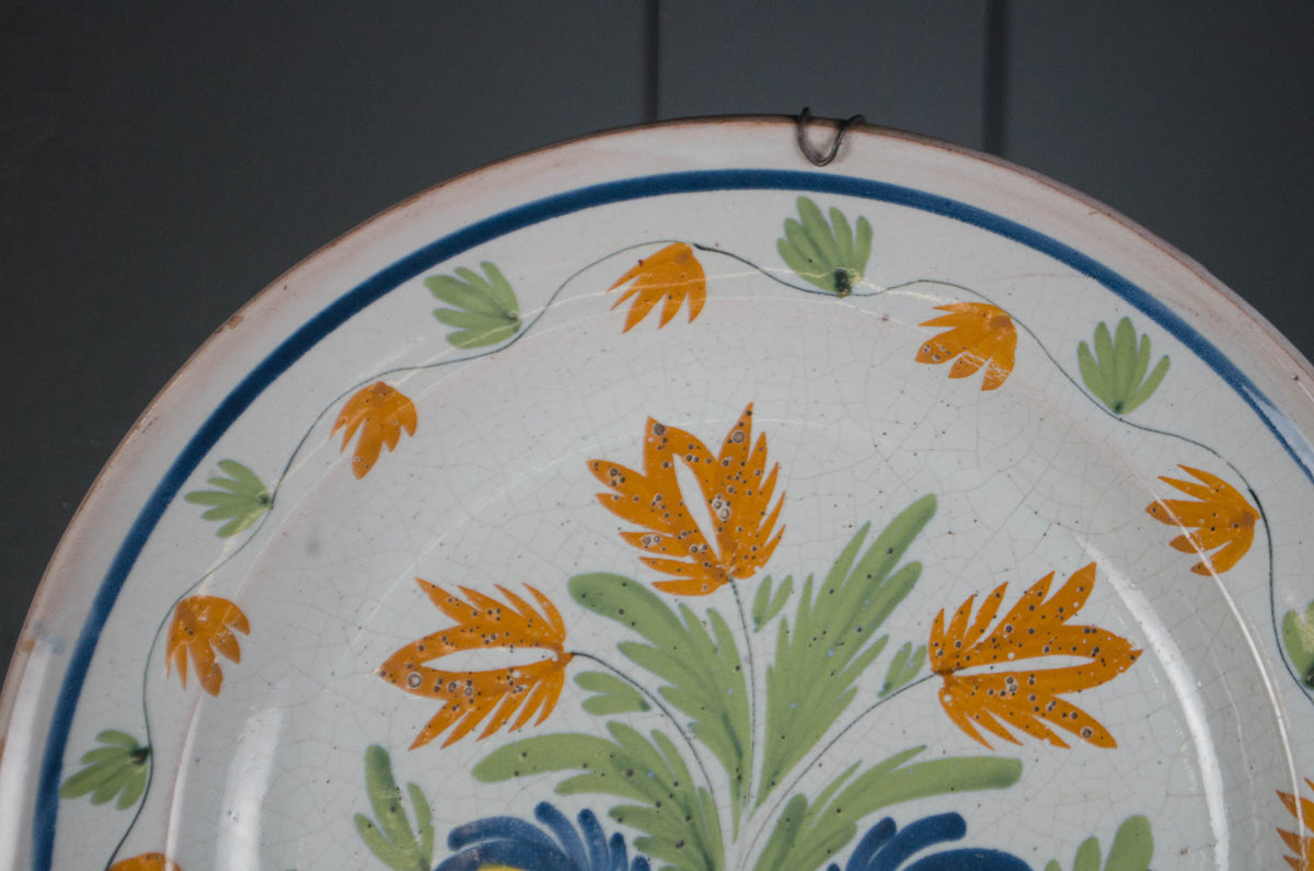 A close-up of a ceramic plate with a hand-painted Mediterranean style floral pattern featuring orange flowers, green leaves, and blue accents on a white background.