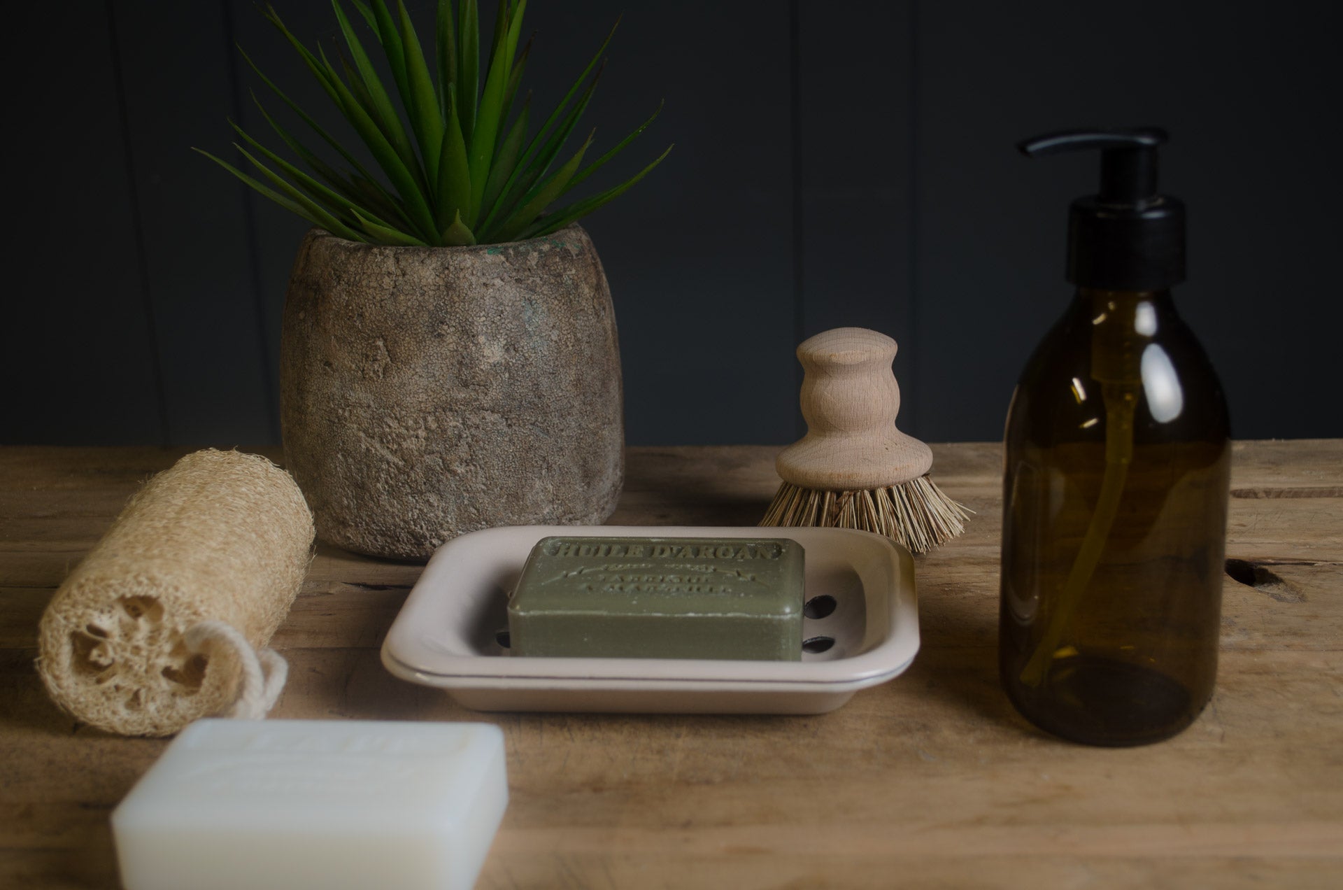 Bar of soap on a soap dish with a bottle, sponge, and plant on a wooden surface. in a framhouse kitchen