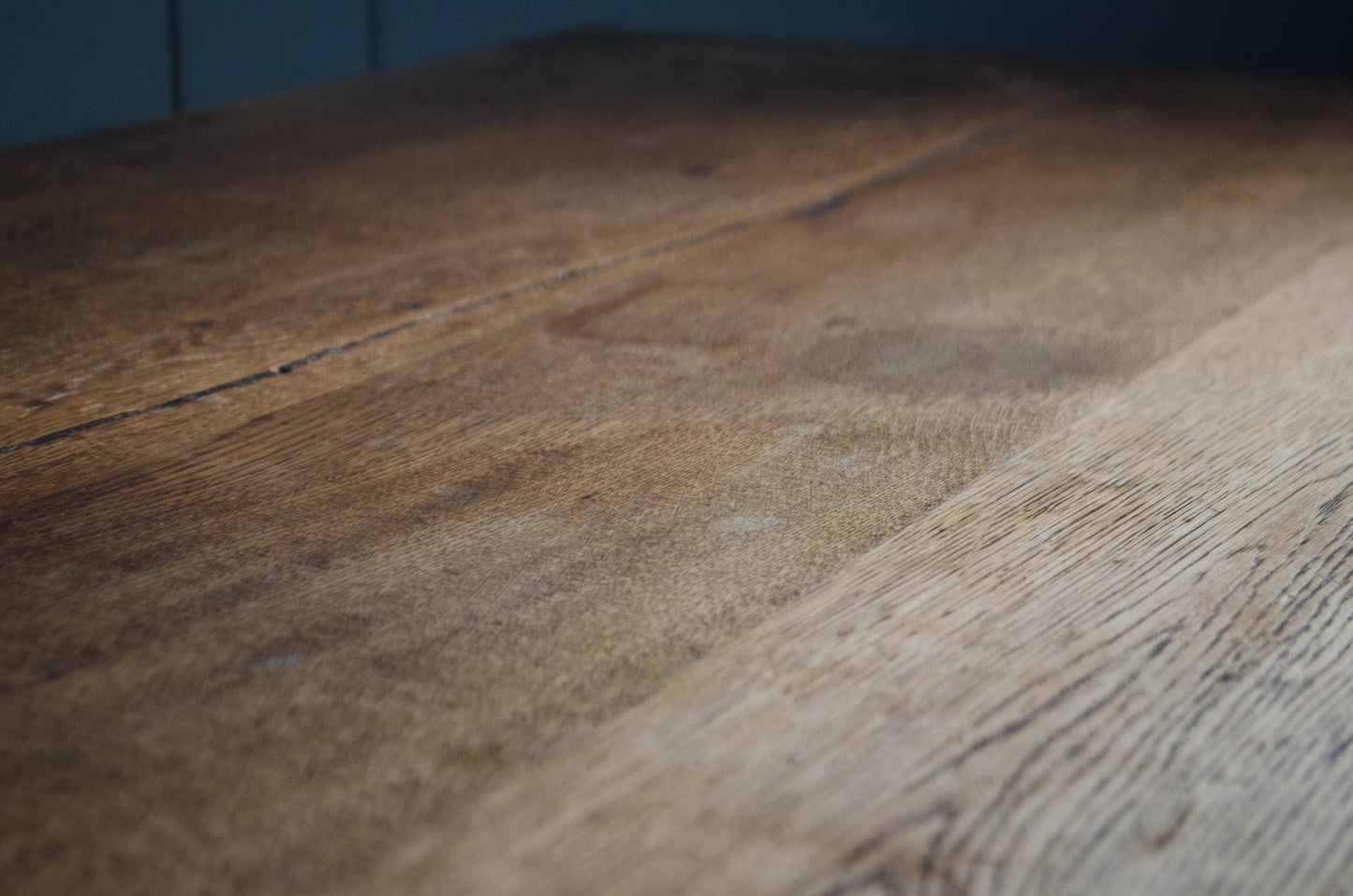 A close-up view of a rustic wooden surface with visible grain, knots, and natural tonal variation, photographed indoors against a dark blue background.