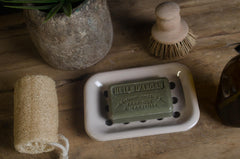 Bar of french soap in a white dish with a wooden brush and sponge on a wooden surface