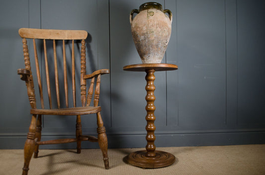 A rustic wooden spindle-back chair sits beside a turned wooden pedestal table holding a large distressed ceramic vase, set against a dark paneled wall.
