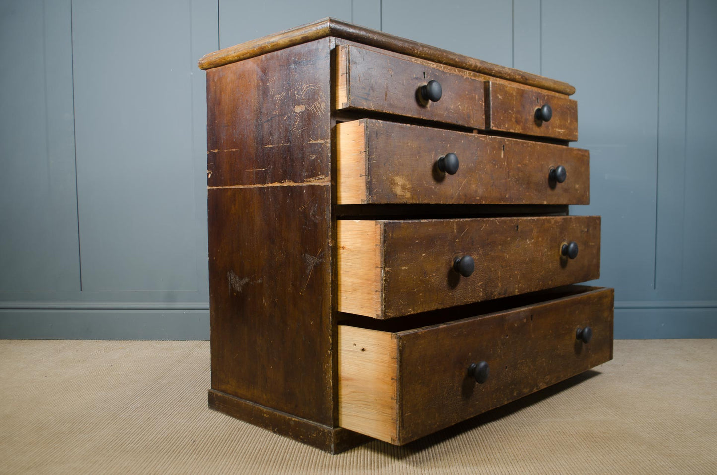A vintage wooden chest of drawers with five drawers, featuring a dark brown finish with lighter natural wood edges and round black knobs, set against a blue paneled wall and beige carpet.