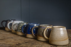 Row of hand thrown pottery ceramic mugs made in Devon with different colors on a wooden surface against a dark background