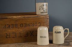Three ceramic containers labeled 'Sugar', 'Flour', and a larger container on a wooden surface with a blue background.