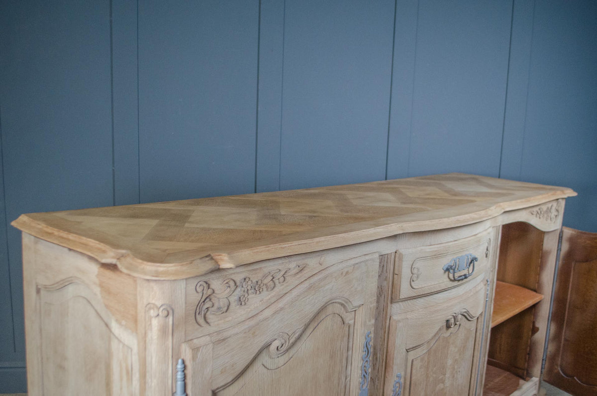 
A light natural wood French-style sideboard cabinet with carved details and a herringbone wood top, set against a blue paneled wall.