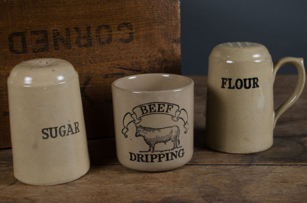 Three ceramic containers labeled 'Sugar', 'Beef Dripping', and 'Flour' on a wooden surface with a wooden box in the background.