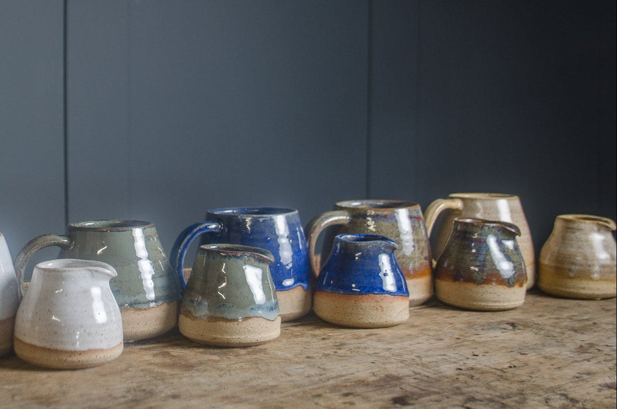 Row of ceramic handmade mini milk jugs in green & mugs in various colours on a wooden surface with a dark grey background