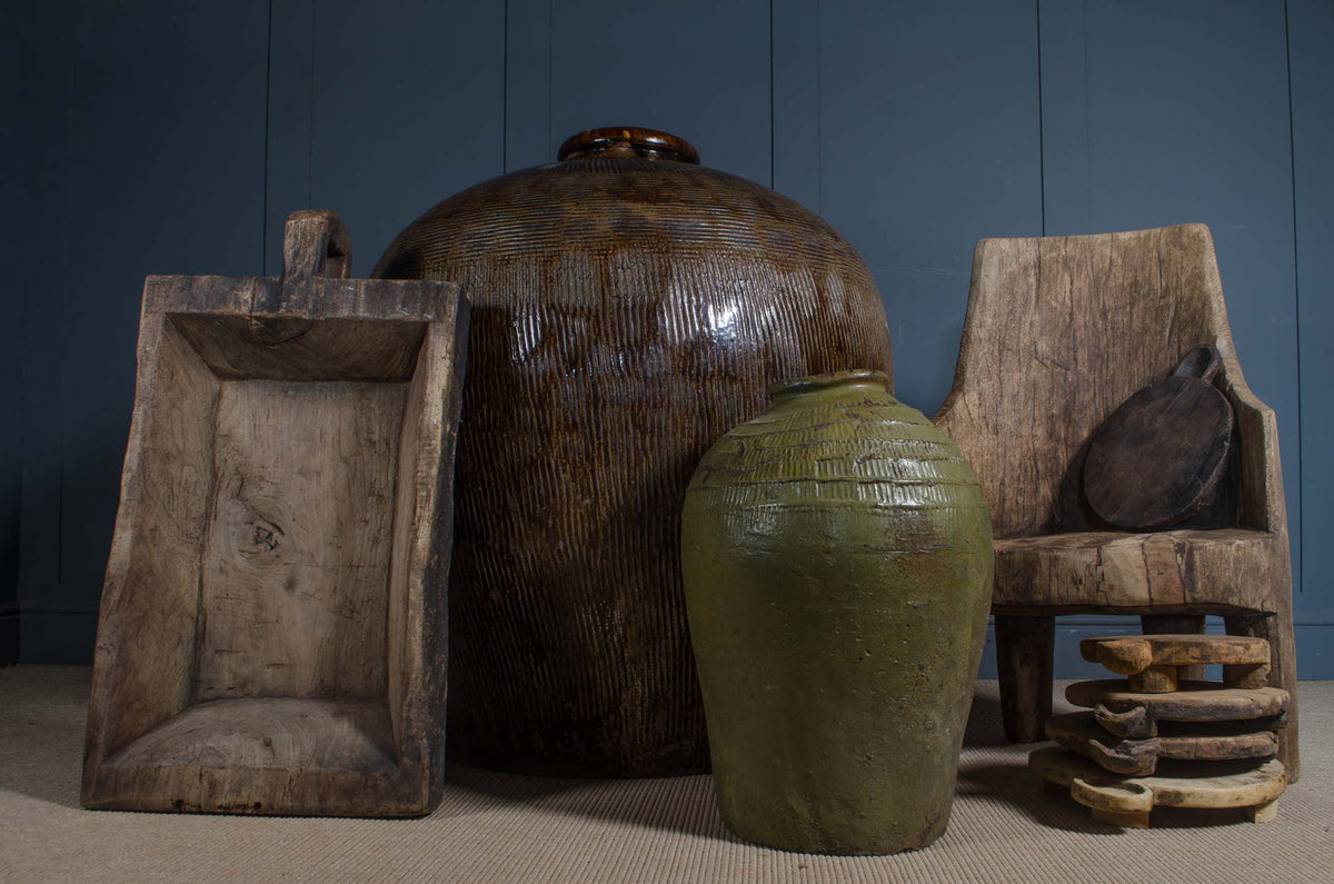 
A collection of rustic vintage objects including a large brown glazed ceramic jar, a smaller green glazed ceramic jar, a carved wooden chair, a wooden scoop, and a stack of small wooden bowls, set against a dark blue paneled wall.