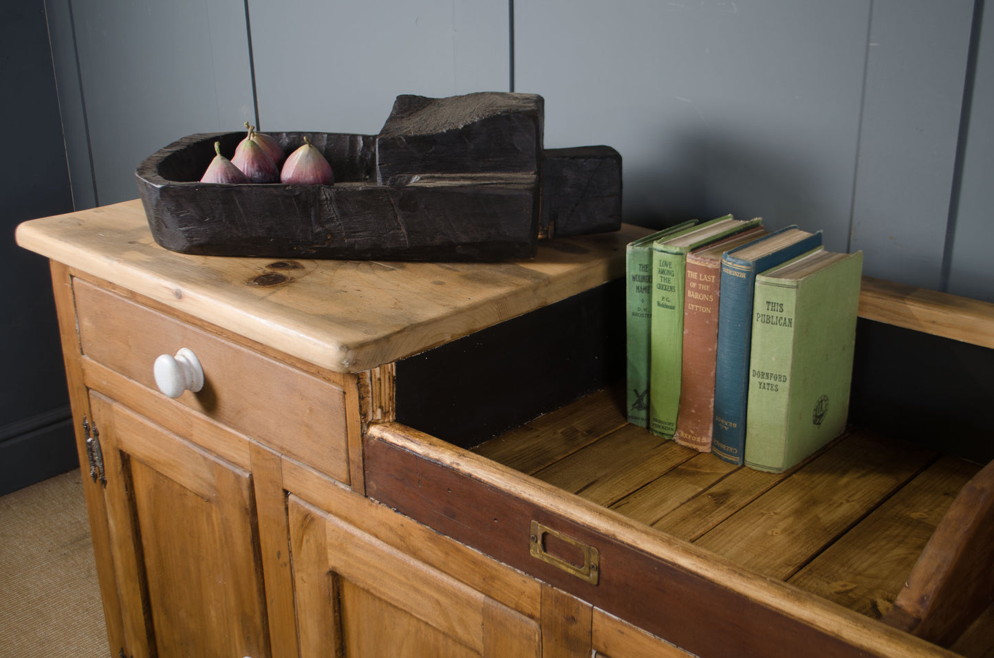 A rustic wooden sideboard cabinet with a light natural wood top, dark brown drawer fronts, and an open shelf holding three vintage books and a dark carved wooden object with pears, set against a dark paneled wall.