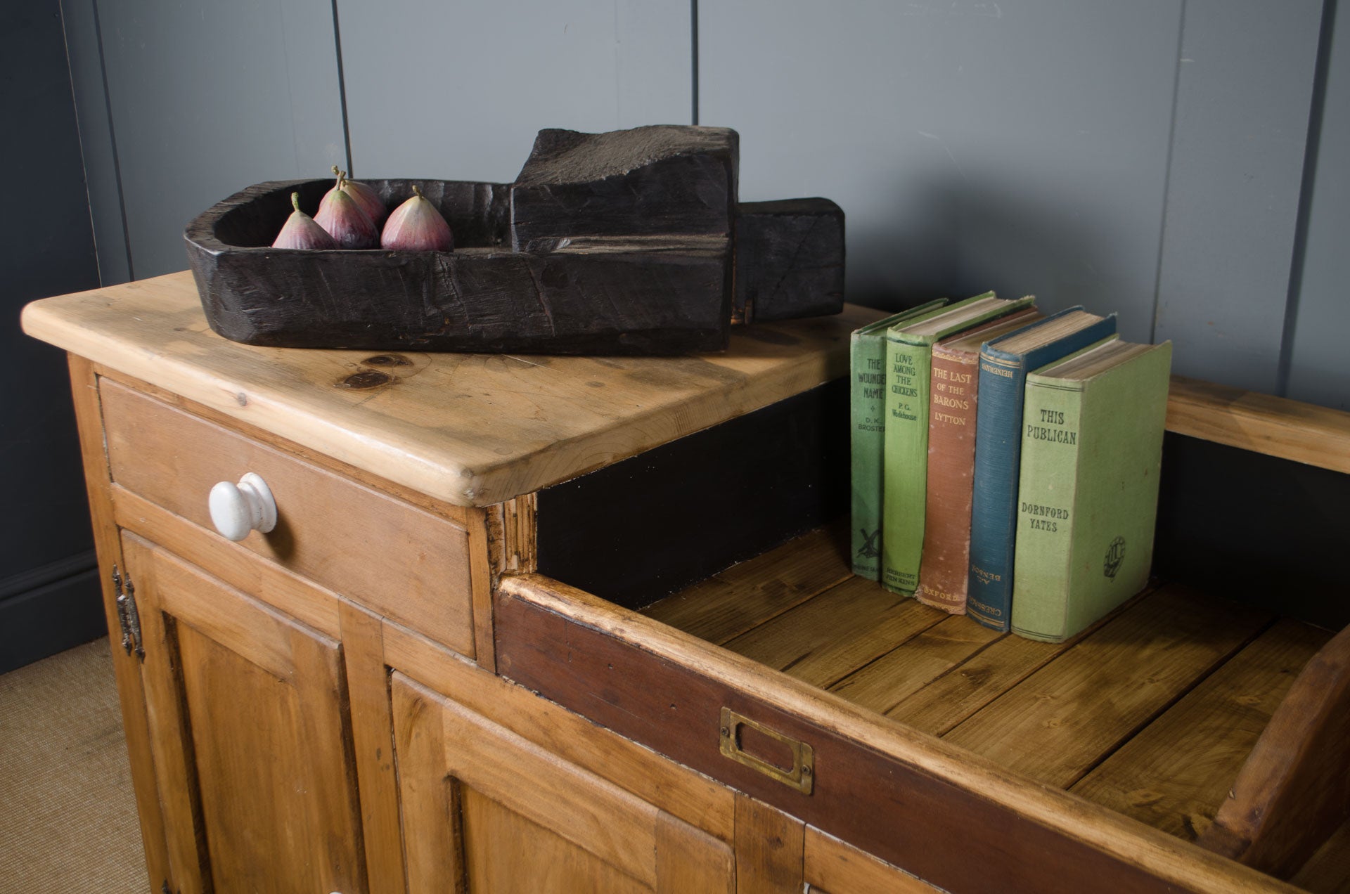 
A rustic wooden sideboard cabinet with a light natural wood top, dark brown drawer fronts, and an open shelf holding three vintage books and a dark carved wooden object with pears, set against a dark paneled wall.