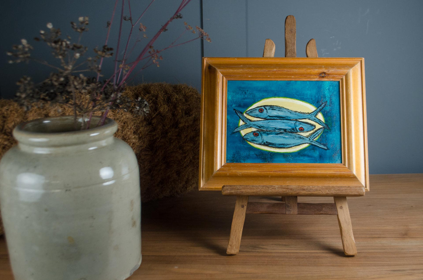 A small framed painting of three blue fish on a yellow background, displayed on a wooden easel stand on a wooden table next to a ceramic jar.