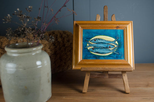 A small framed painting of three blue fish on a yellow background, displayed on a wooden easel stand on a wooden table next to a ceramic jar.
