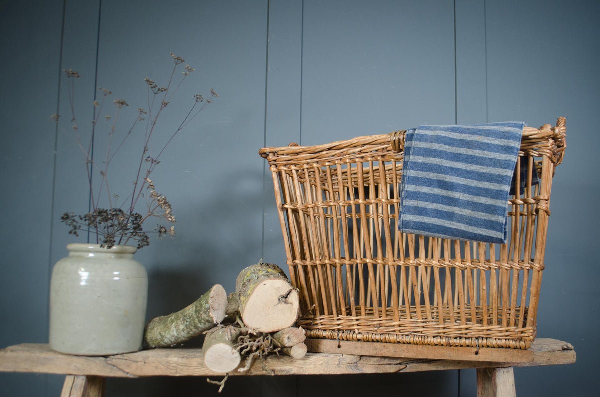 
A natural wicker laundry basket with a blue and white striped towel draped over the side, styled on a rustic wooden bench next to a ceramic vase with dried stems and cut wood logs against a blue paneled wall.