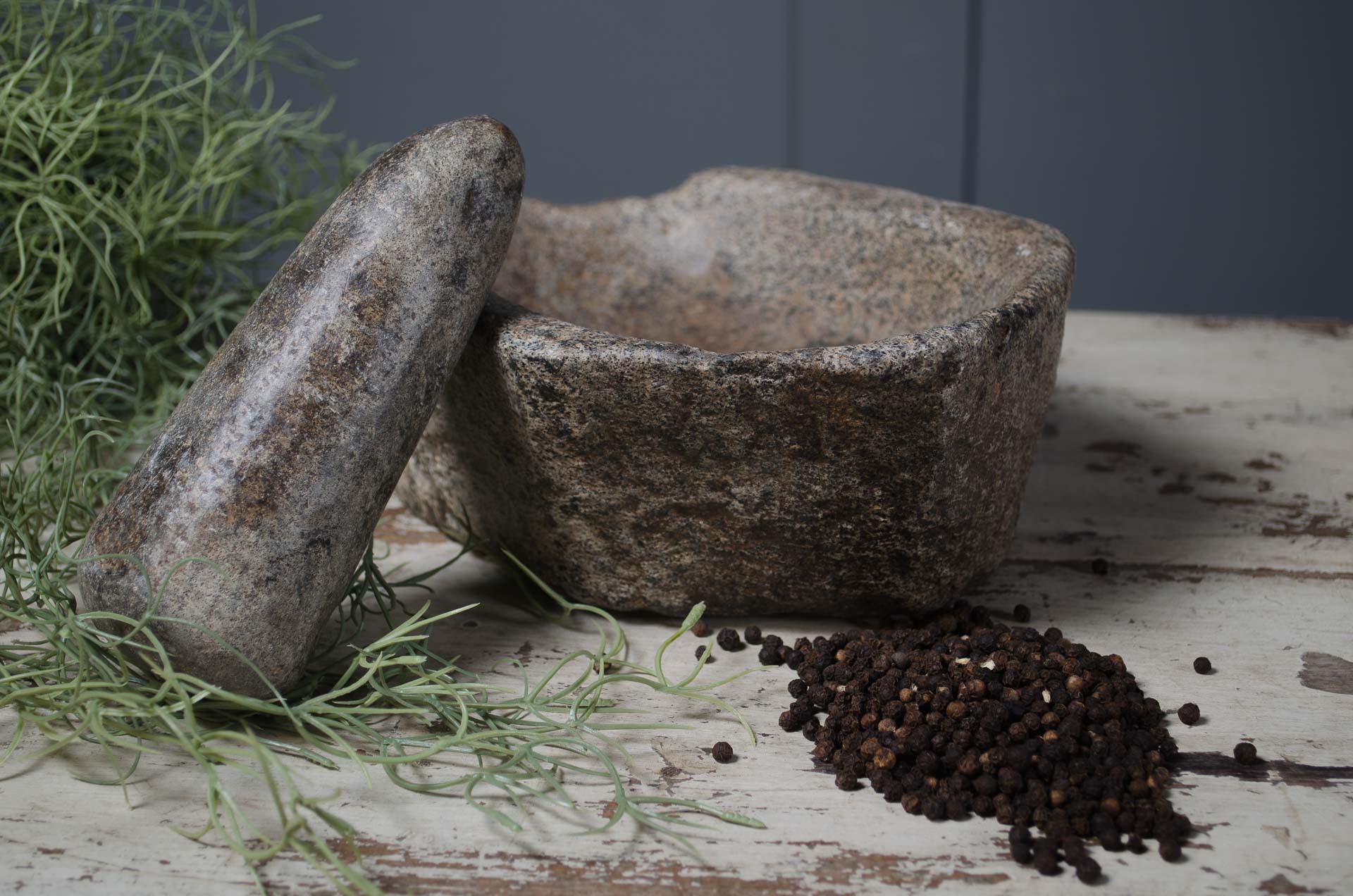 Two stone mortars and a pile of black peppercorns on a rustic wooden surface with greenery.