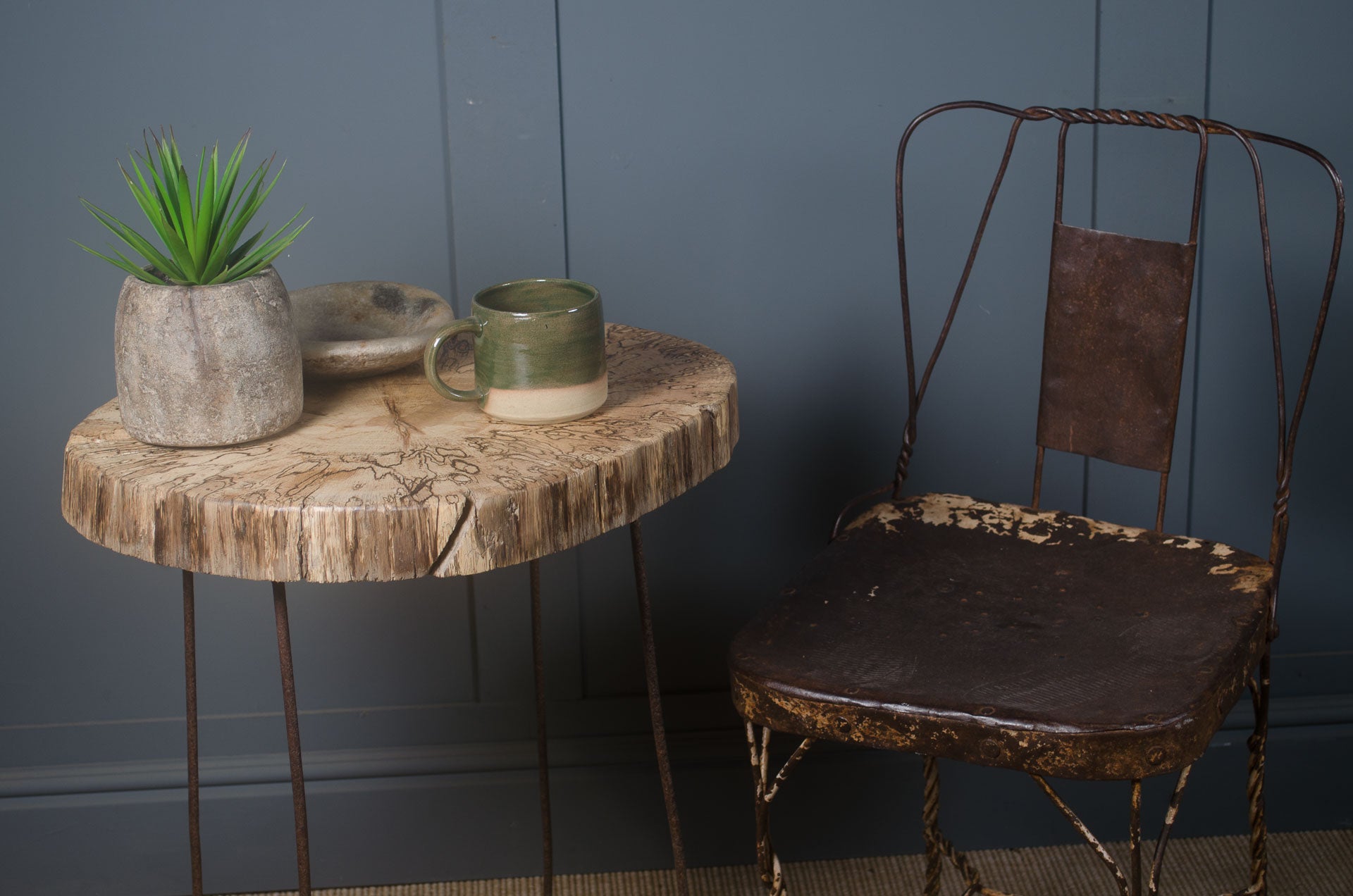Small round wooden table with a plant and mug next to an old metal chair against a blue wall.