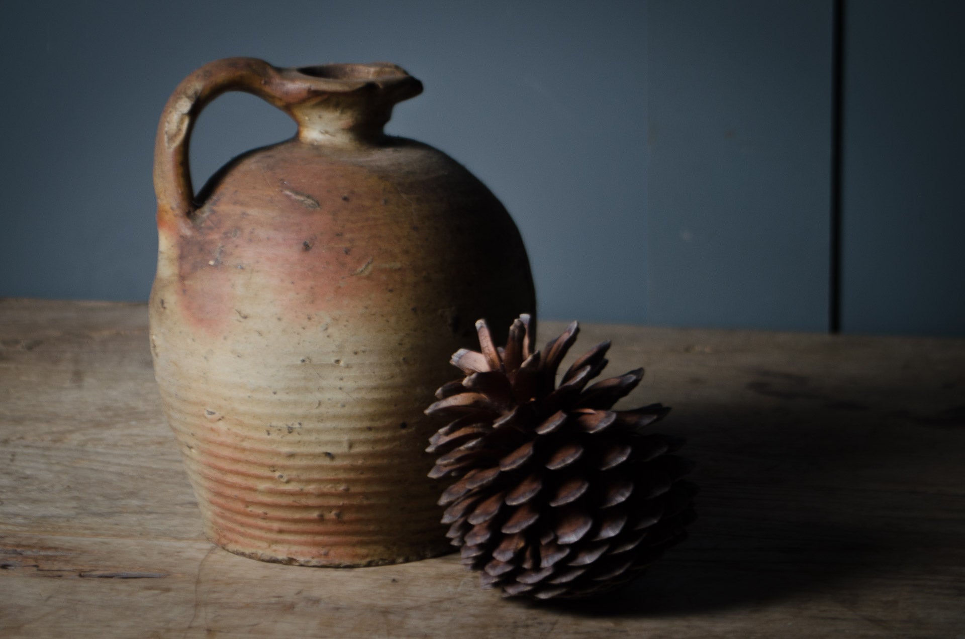Stoneware jar and pine cone on a wooden surface with a dark background. french oil jug