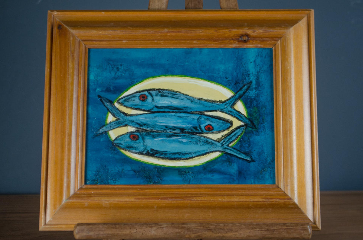 
A framed painting of three blue fish on a textured blue background, displayed on a wooden easel against a dark gray wall.