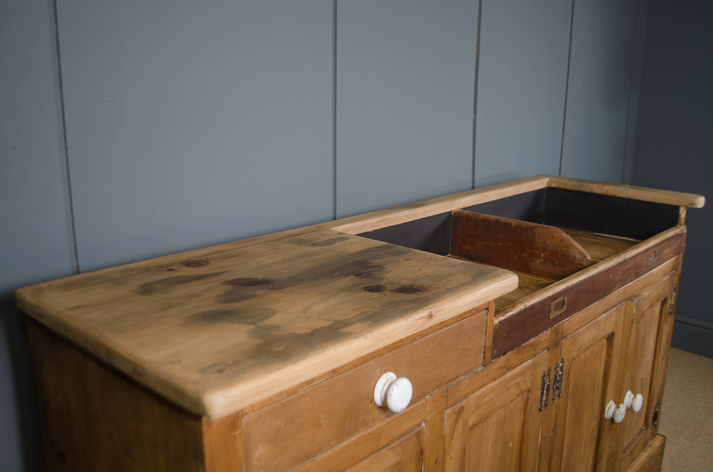 A vintage wooden kitchen sink cabinet with a large wooden countertop, a built-in sink, and white knobs, set against a dark gray paneled wall.