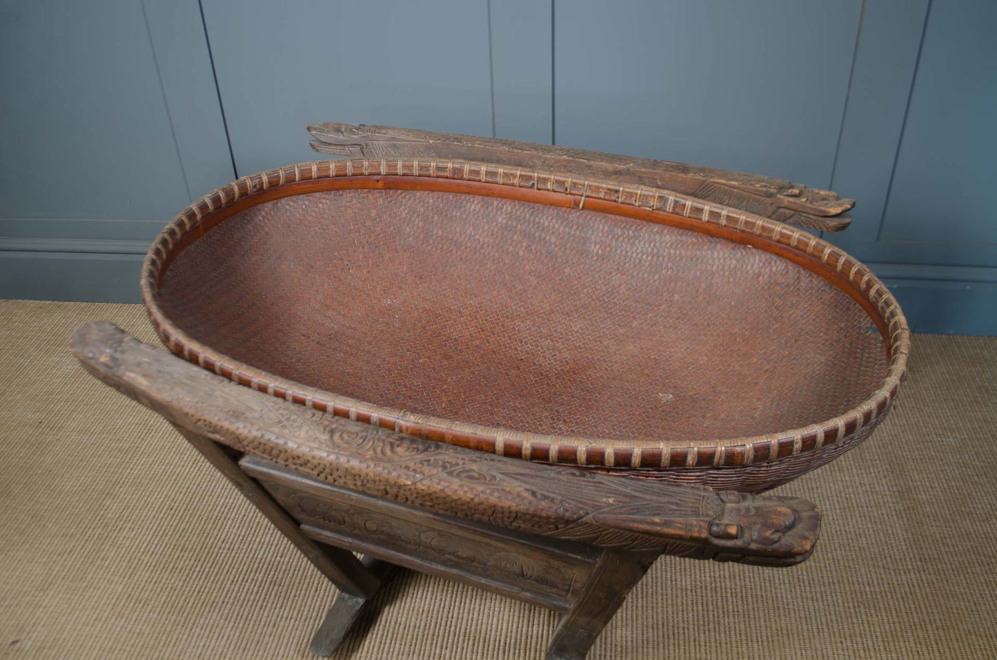 An antique oval wooden basket with a woven brown inner lining, displayed on a rustic wooden stand against a blue paneled wall.