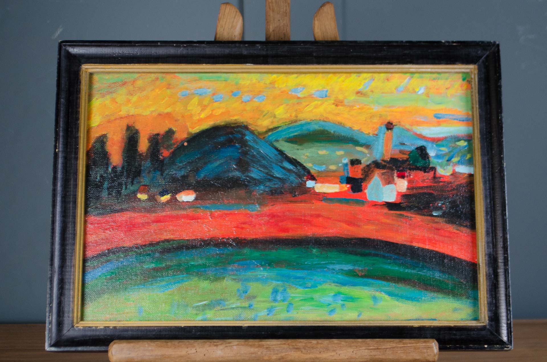 
A framed landscape oil painting on canvas, displayed on a wooden easel against a dark gray wall. The painting features a vibrant, impressionistic scene with a bright yellow sky, dark blue hills, a red field, and green foreground, enclosed in a black frame.