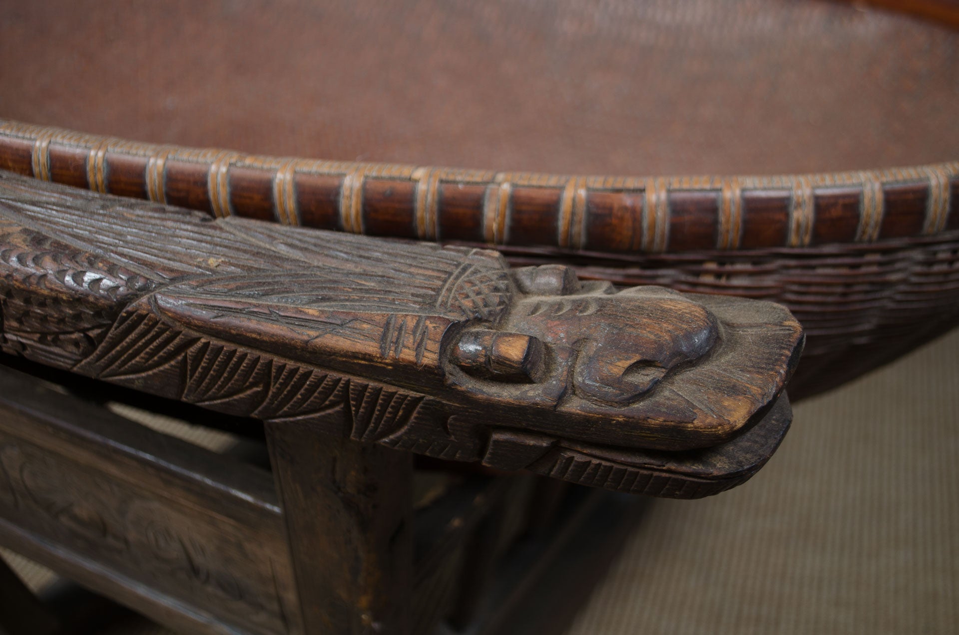 
Close-up of a carved wooden table corner with a detailed dragon head topper and a woven basket or tray rim in the background.