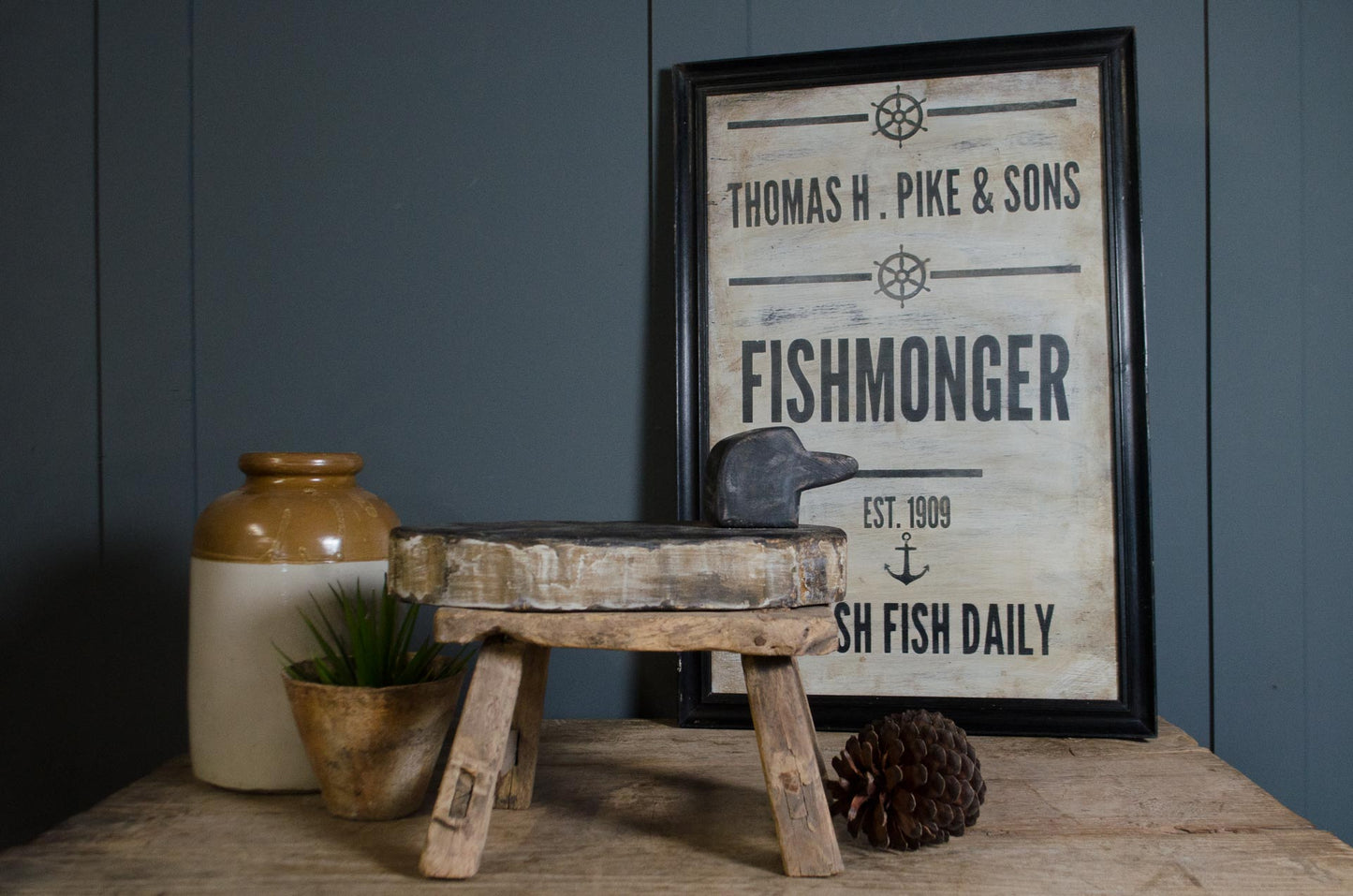 A framed vintage-style fishmonger sign with black text on a distressed cream background, styled on a wooden table with a rustic stool, ceramic jar, small plant, and pinecone against a dark blue paneled wall.