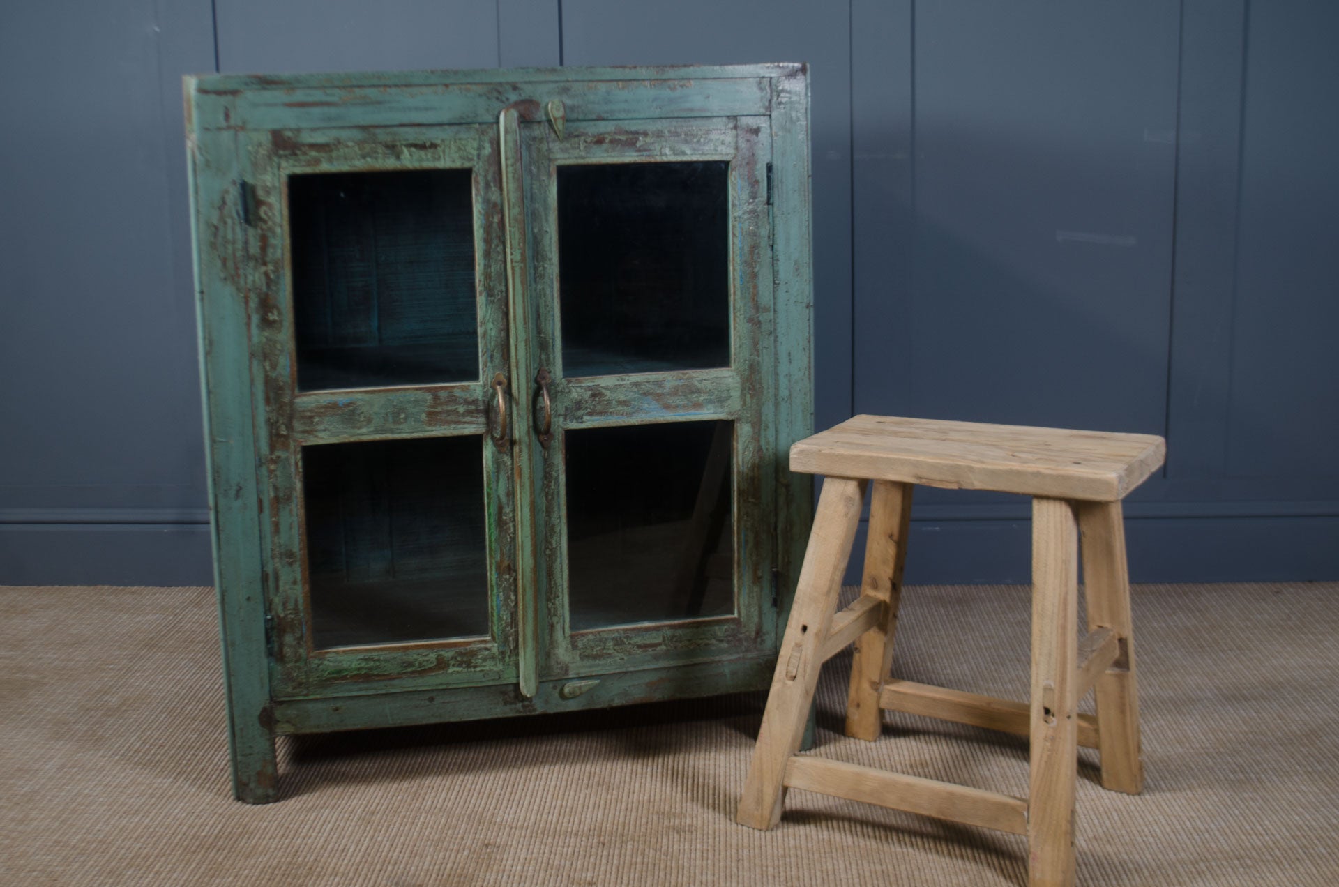 
Vintage-style cabinet with green patina and a wooden stool against a blue wall.