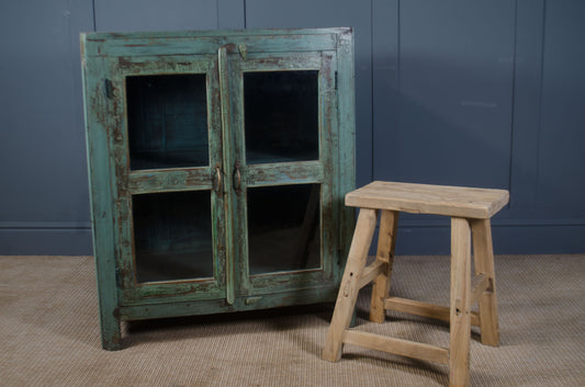 Vintage-style cabinet with green patina and a wooden stool against a blue wall.