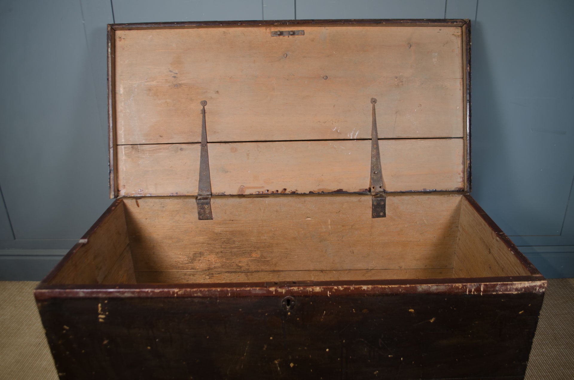 
An antique wooden storage chest with its lid open, showing a dark brown exterior and a light natural wood interior, set against a blue paneled wall.
