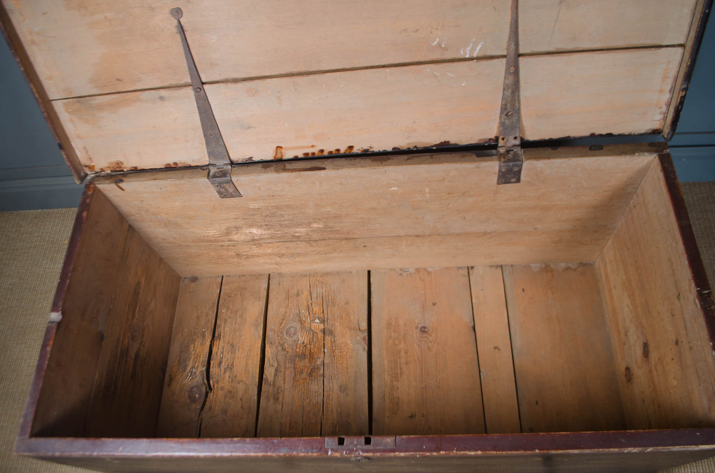 An open vintage wooden storage chest or trunk with a slatted wooden interior and dark metal hinges, photographed indoors against a blue wall and beige floor.