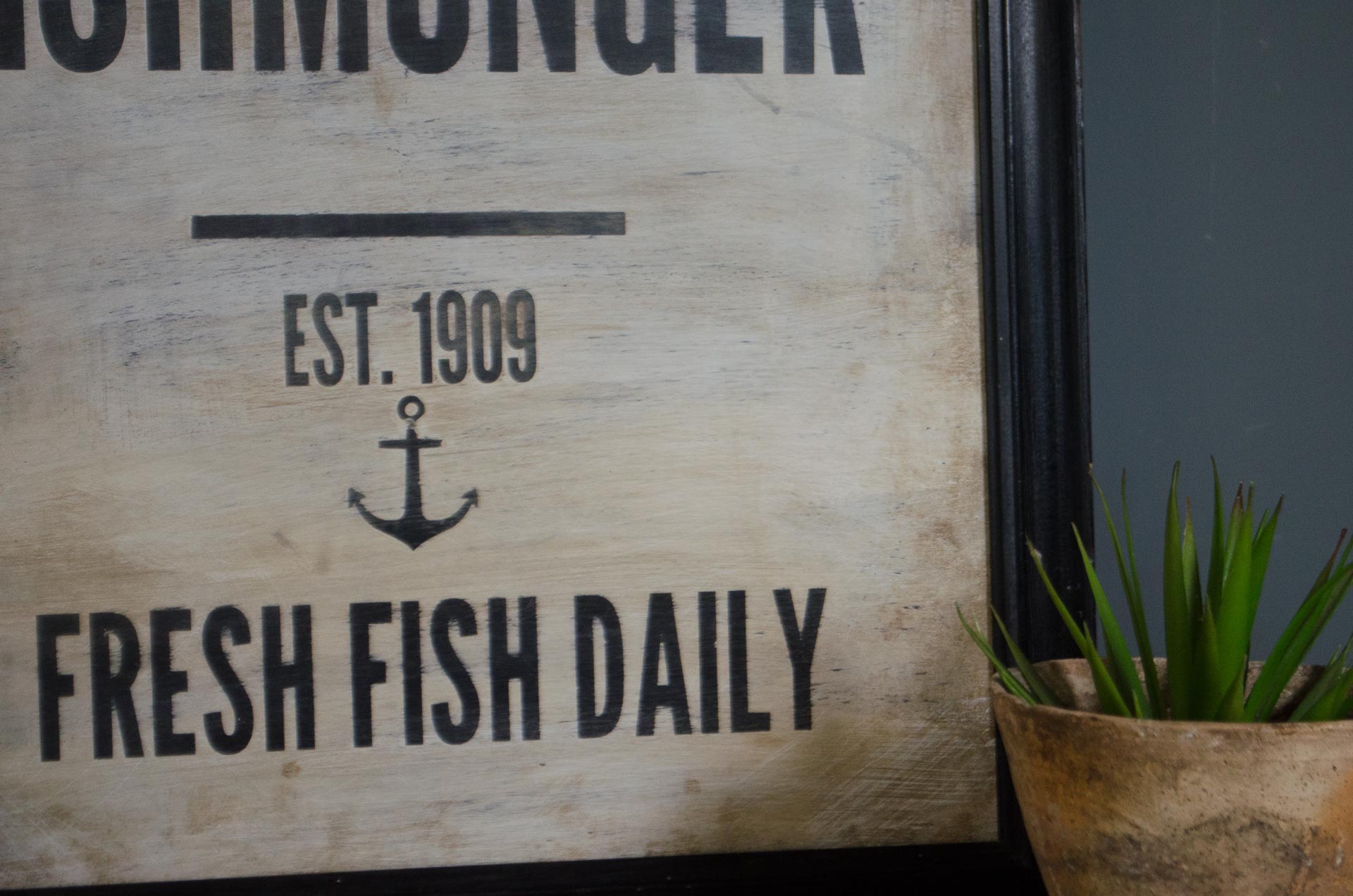 
A framed rustic wooden sign with black text reading 'NIMONDER', 'EST. 1909', and 'FRESH FISH DAILY', shown next to a small potted green plant.