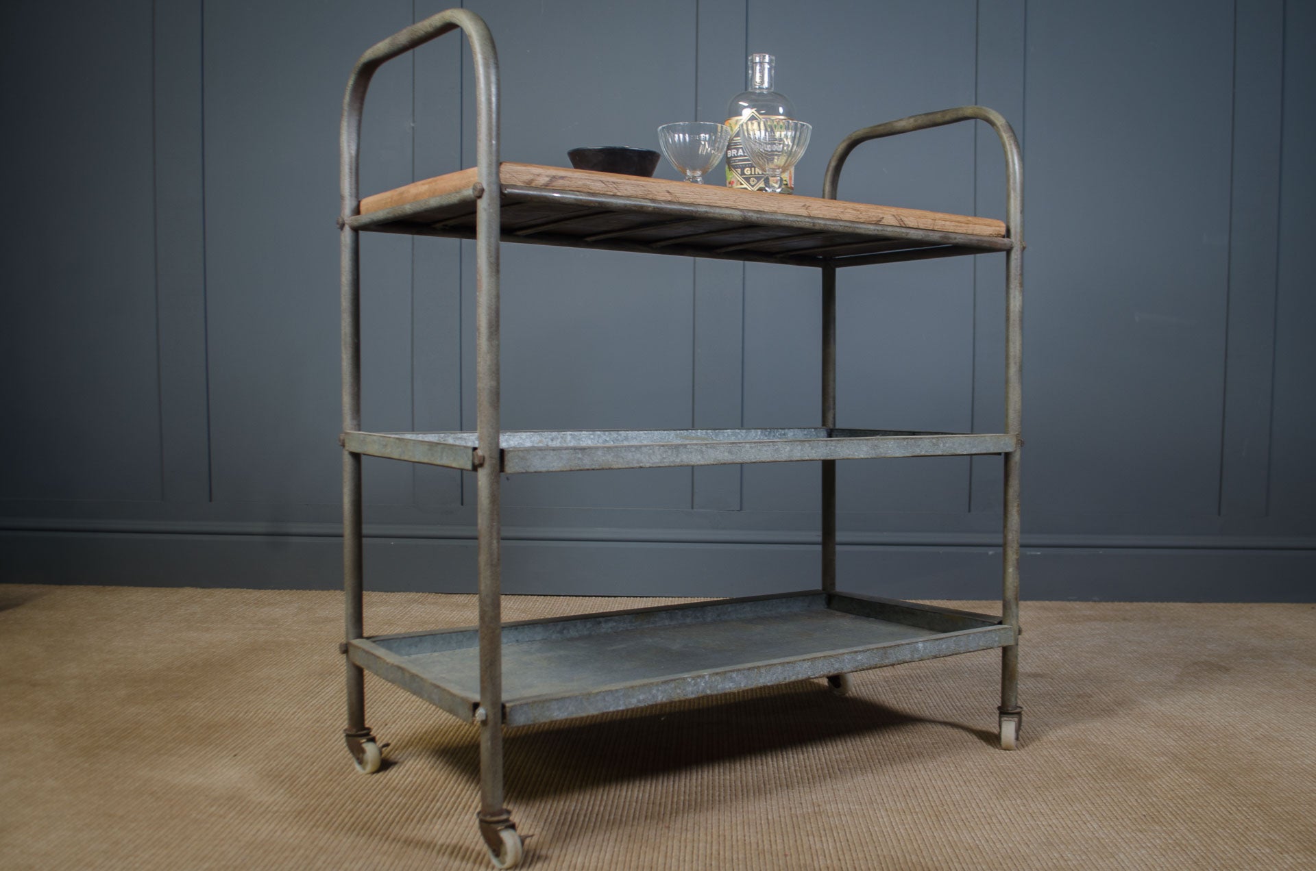 Vintage metal serving cart with wooden top against a gray wall.