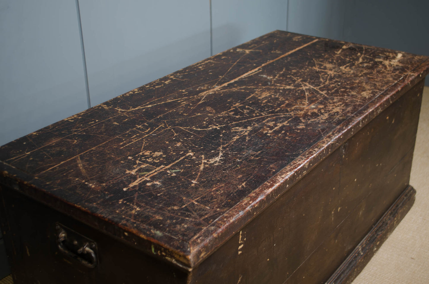 A dark brown wooden storage chest or trunk with a heavily worn, scratched, and scuffed wooden lid, set against a gray paneled wall.