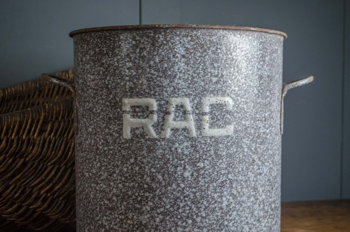 A tall speckled gray enamel storage canister with metal side handles and the letters 'RAC' embossed on the front, set against a dark gray background with a wicker basket partially visible on the left.