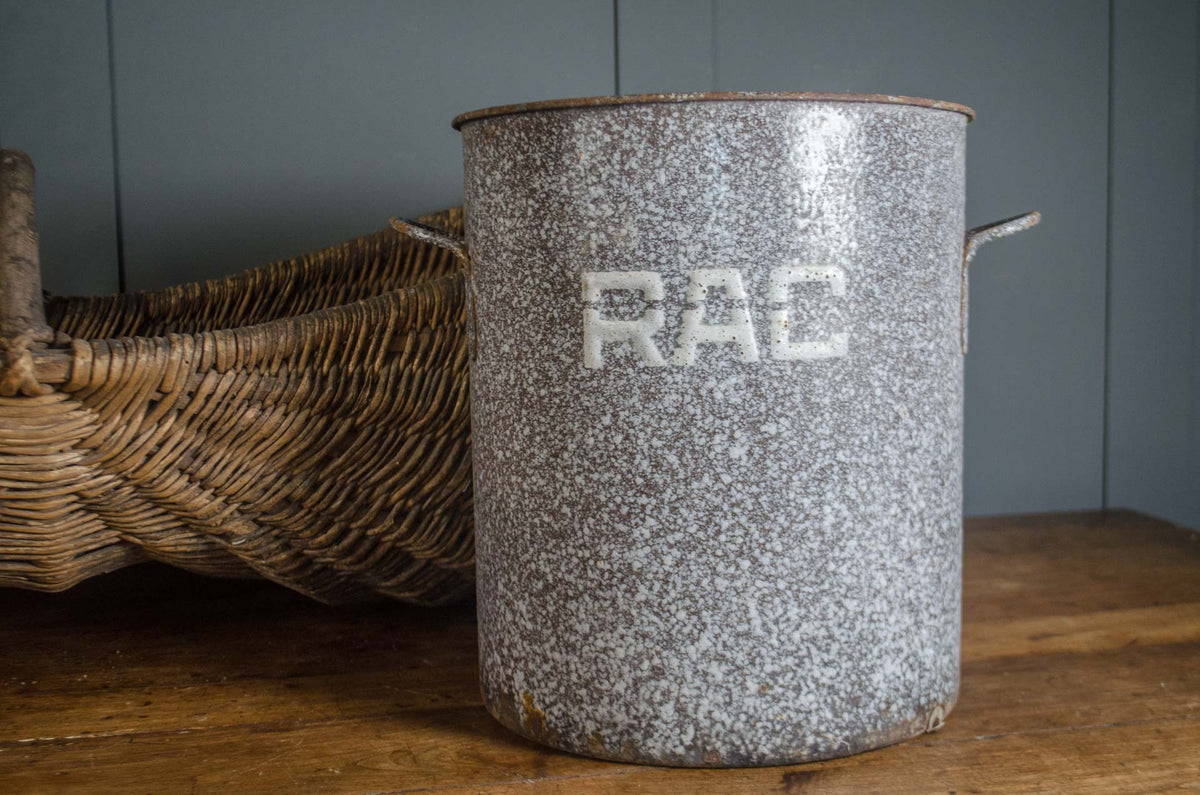 
A vintage speckled gray enamel canister with white 'RAC' lettering, sitting on a rustic wooden table next to a woven wicker basket.