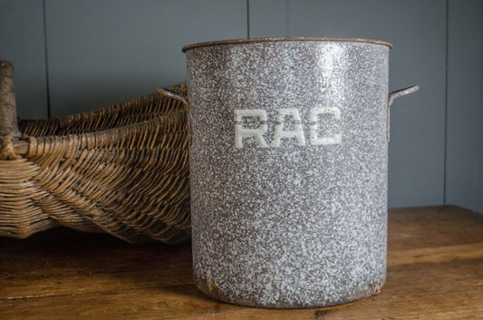 A vintage speckled gray enamel canister with white 'RAC' lettering, sitting on a rustic wooden table next to a woven wicker basket.