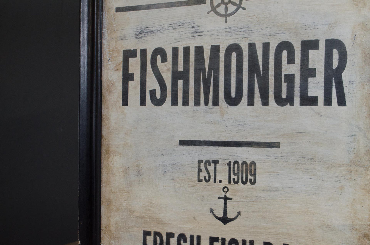 A close-up of a rustic wooden sign with distressed beige wood grain and black printed text reading 'FISHMONGER', 'EST. 1909', and an anchor symbol.