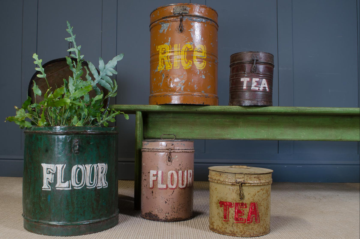 A styled collection of vintage metal canisters and buckets in green, rust orange, and pink, some decorated with white, yellow, and red lettering, arranged on a green bench against a dark gray paneled wall.