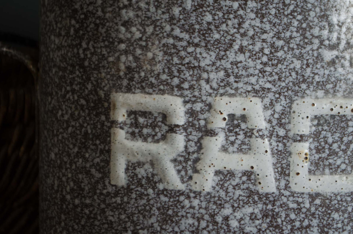 Close-up of a speckled ceramic mug or container with a brown and white marbled glaze and the letters 'RAC' in white raised on the front.