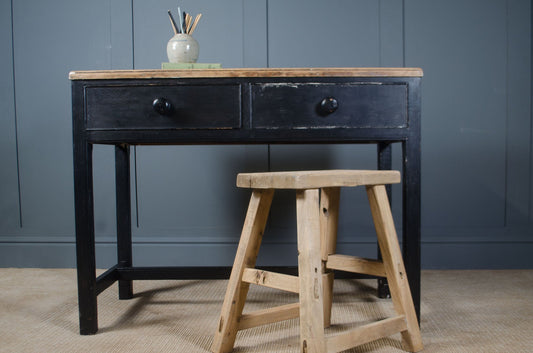 Black wooden desk with two drawers and a wooden stool in front against a dark blue wall.