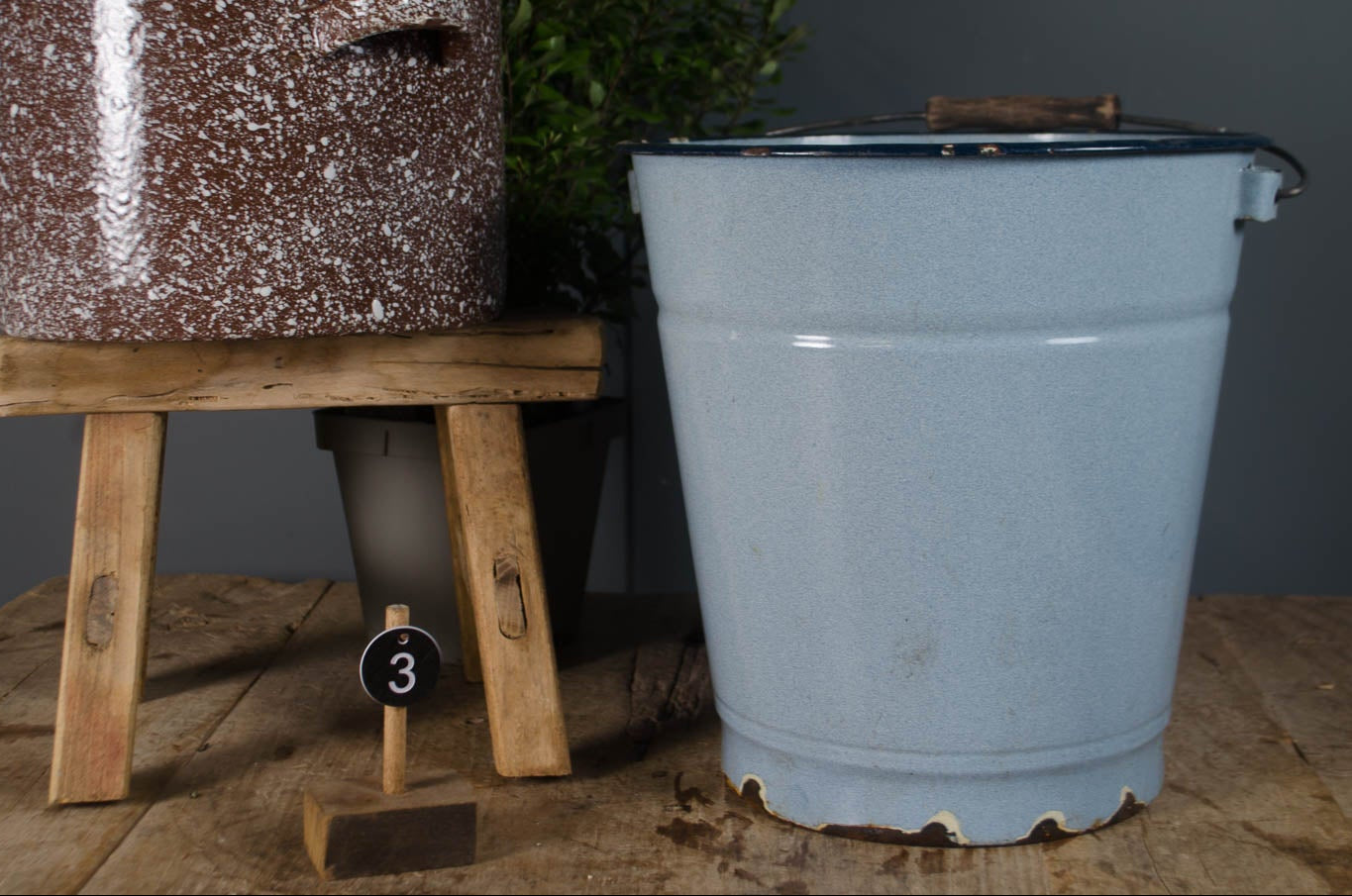 Speckled brown planter with flowers on a wooden stool next to a blue metal bucket against a gray wall.