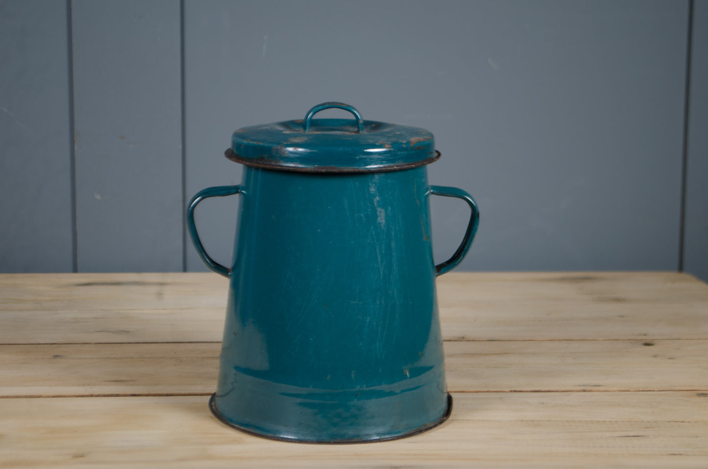 Teal metal bucket with handles on a wooden surface against a gray wall.