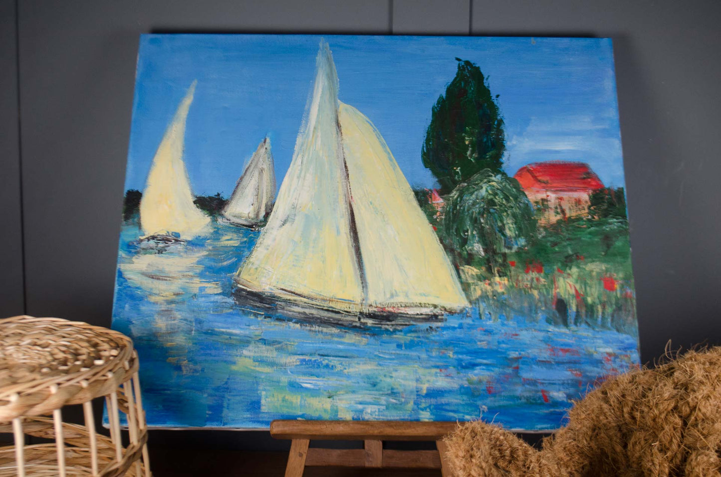 An impressionist-style oil painting of sailboats on blue water, displayed on a wooden easel against a dark gray wall. The painting features a prominent sailboat with cream-colored sails, other boats in the background, a dark green tree, and a house with a red roof.