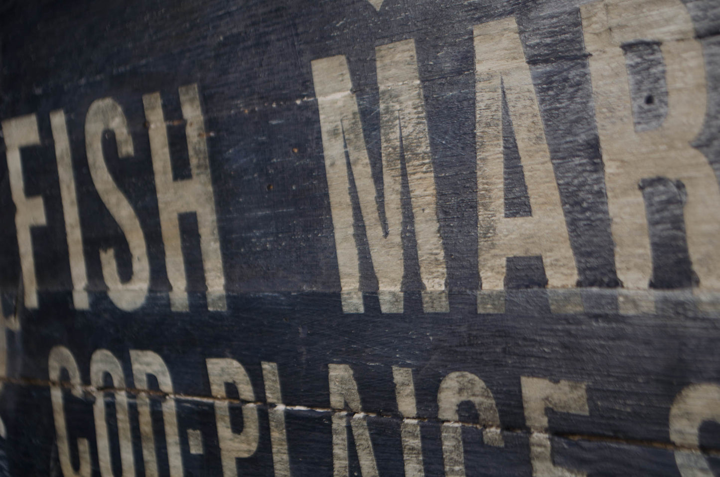A close-up of a weathered wooden sign with distressed cream-colored lettering reading 'FISH MAR' and 'COD-PLINCE' on a dark painted wood surface.