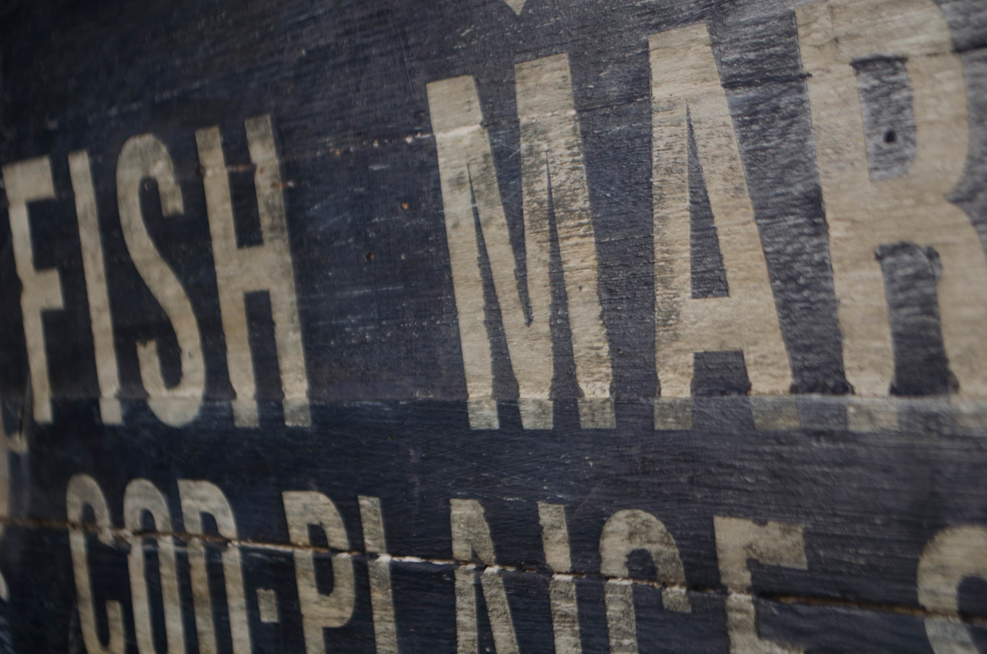 
A close-up of a weathered wooden sign with distressed cream-colored lettering reading 'FISH MAR' and 'COD-PLINCE' on a dark painted wood surface.