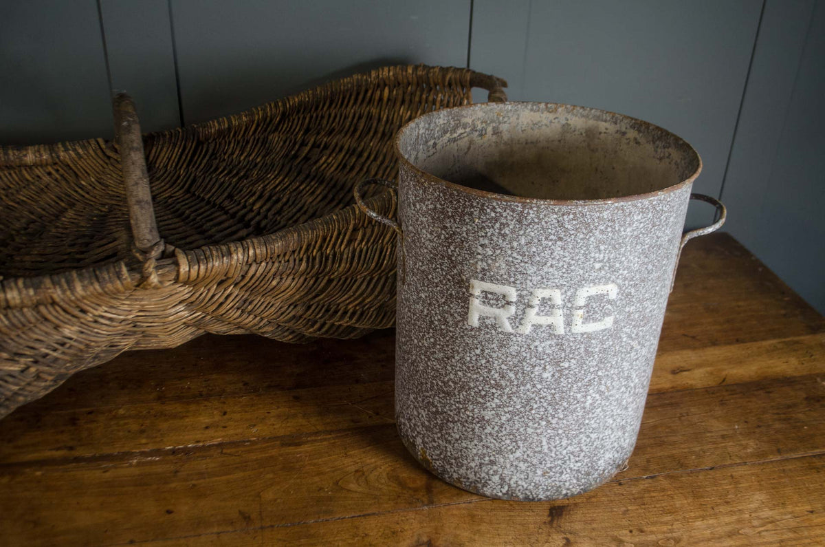 
A speckled gray enamel metal bucket with side handles and the letters 'RAC' painted on the front, sitting on a rustic wooden table next to a woven wicker basket.